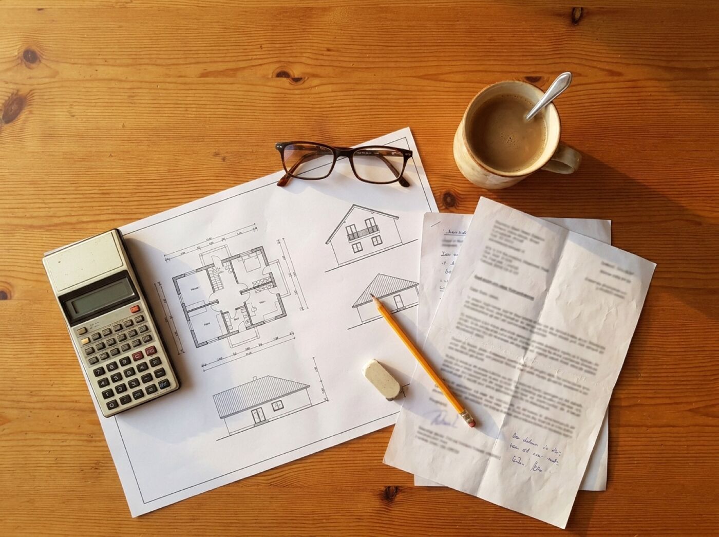Hausgrundriss, Taschenrechner, Lesebrille und Unterlagen auf einem Holztisch bei der Finanzierungsplanung